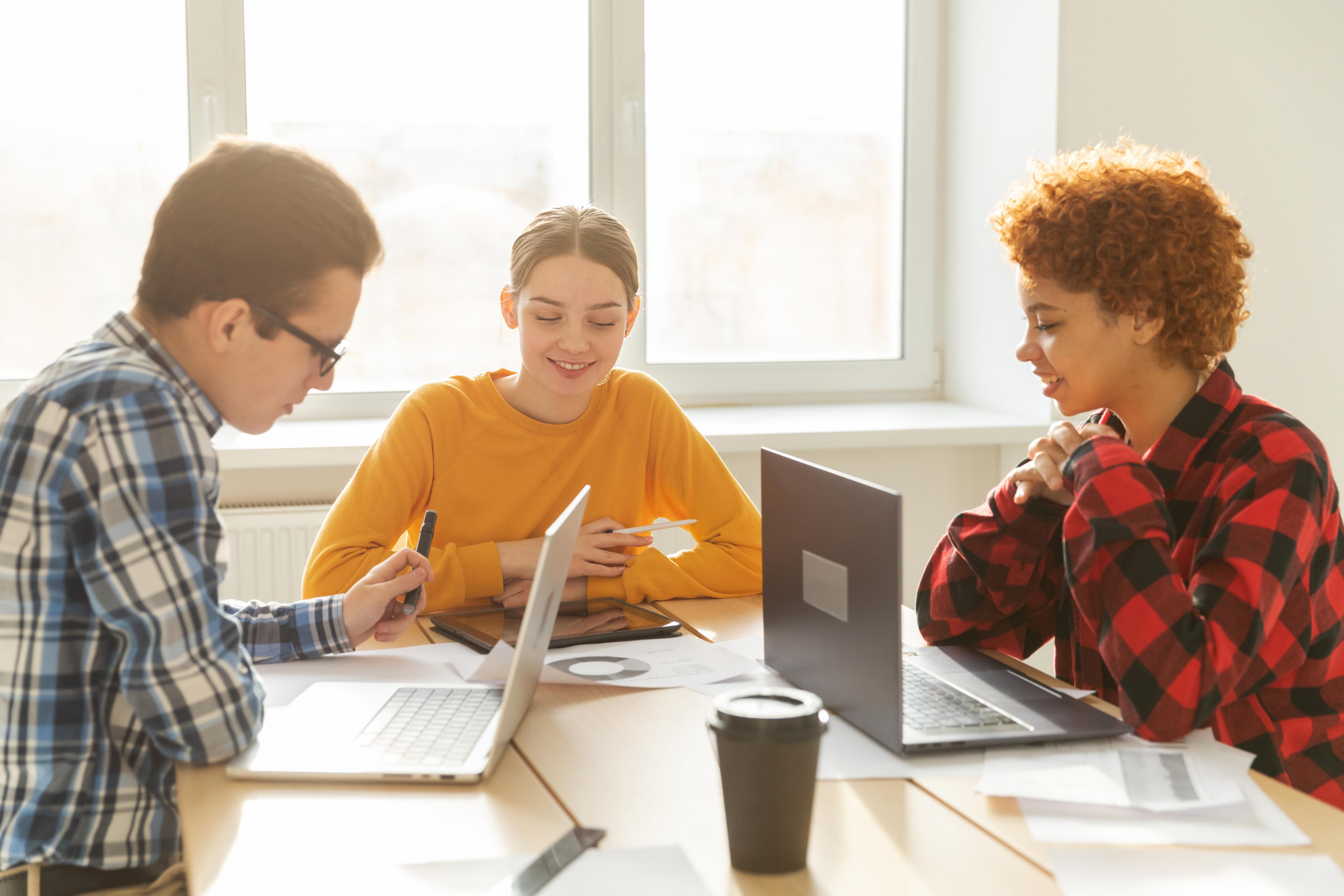 Educators collaborating with students to prepare for the Digital SAT, using laptops and test prep materials in a bright classroom.